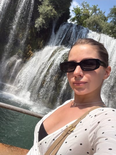 An outdoor, medium shot selfie shows a young woman with brown hair pulled back and wearing dark sunglasses, a white polka-dot top, and a light brown crossbody bag strap. She is looking towards the camera, with a smile. Behind her, a large waterfall cascades down rocks, with lush green trees visible at the top. The water is clear and turquoise, and a metal railing is visible in the foreground. The scene appears to be the Pliva Waterfall in Jajce, Bosnia and Herzegovina, a natural landmark with a historic old town and several mills.