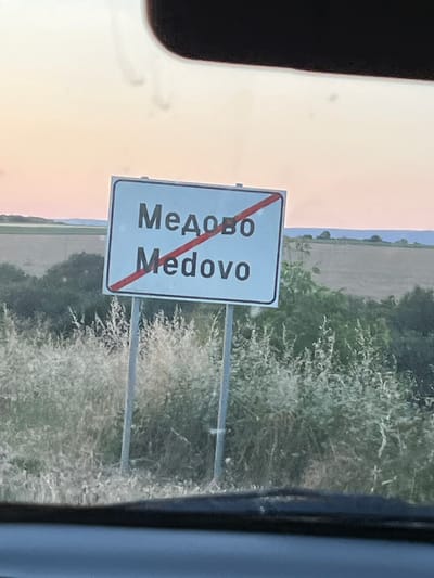 The image shows a road sign with text in both Cyrillic and Latin alphabets, indicating "Медово" and "Medovo" respectively. A red diagonal line crosses both lines of text, signifying the end of a settlement. The sign is mounted on two metal posts and is situated in a rural setting with fields and vegetation in the background. The sky is visible above with a pale, orange hue, suggesting either sunrise or sunset. The foreground is blurred, indicating the photo was taken from a moving vehicle.