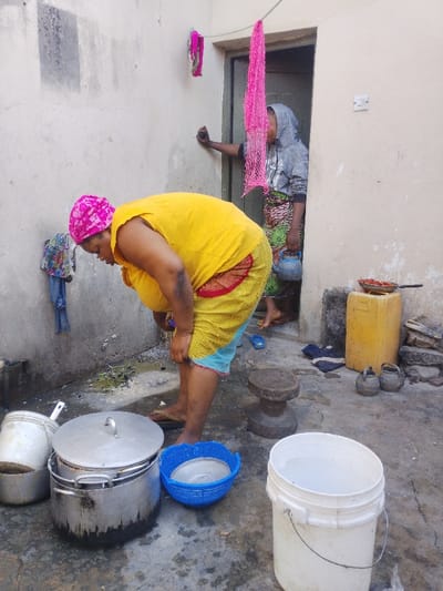 A woman in a yellow shirt and pink head covering is washing items near a wall outside. A variety of pots, pans, and a blue colander are on the ground. Behind her, a woman in a hoodie and patterned skirt holds a blue kettle, leaning against the doorway. Pink laundry hangs above the door. A yellow container and small pots are near the entrance. The space appears to be an outdoor courtyard or service area.