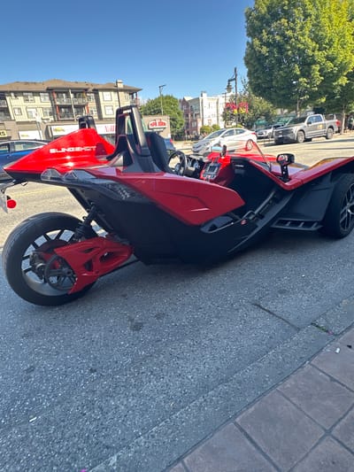 A red Polaris Slingshot is parked on a paved street in front of buildings with businesses including "Papa John's". The vehicle is angled towards the right side of the frame, with its rear wheel and part of its suspension system prominently displayed on the left. The sky is clear and blue, and trees with green foliage are visible in the background. The scene appears to be in a commercial area, likely in the city of Port Coquitlam, Canada, given the presence of a Canadian flag sticker on the vehicle's side panel.