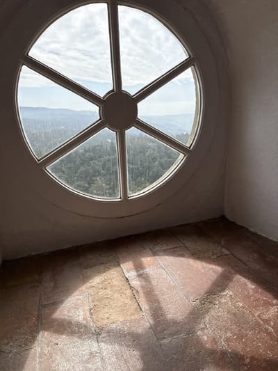 A round window with six panes of glass, set into a white plaster wall, overlooks a landscape of trees and hills under a partly cloudy sky. The floor is tiled with rectangular earth-toned tiles, and shadows from the window frames are cast across the floor. This is a view from Quinta da Regaleira in Sintra, Portugal.