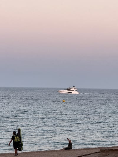 The image captures a scene at Platja D'aro, Spain, featuring a man in the foreground with a black and green backpack. He is positioned on what appears to be a rocky cliffside or pier, looking out towards the ocean. In the middle ground, a white catamaran sails on a calm, blue sea under a bright sky, suggesting it's daytime with clear weather. Further in the distance, another person is visible on the same rocky outcrop as the foreground subject, seemingly observing the catamaran or the scenery. The overall mood is tranquil and recreational, with the vastness of the ocean and the clear sky contributing to a sense of peace. The rock formation the people are standing on could be part of the coastline of Platja D'aro, a popular tourist destination known for its rocky coves and beaches. There is no visible text in the image.