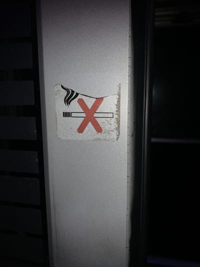 A worn, torn no-smoking sticker is adhered to a gray surface. The sticker depicts a cigarette with a red cross over it. Smoke plumes emerge from the cigarette's lit end. The sticker's edges are frayed, and a portion is missing near the smoke. A black horizontal bar runs across the top of the image. Below the sticker, a series of black vertical rectangles creates a grid-like pattern. The overall lighting is dim.