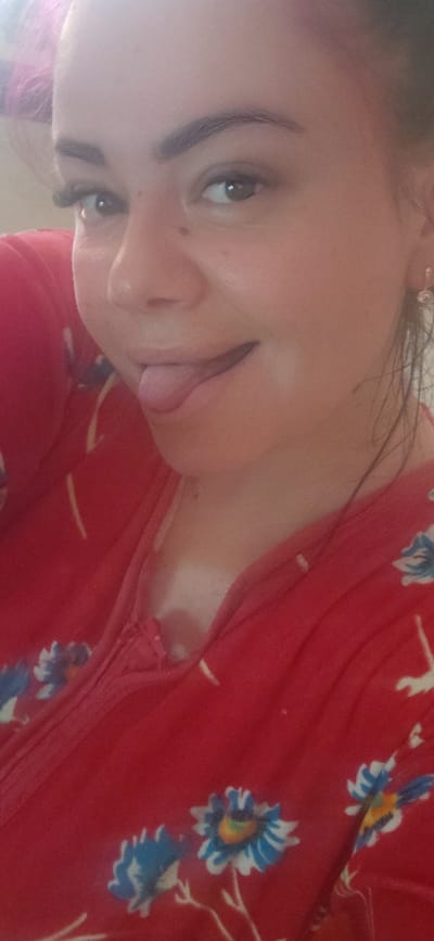 A close-up, selfie-style photograph shows a woman with light-colored skin and dark eyebrows. She is wearing a red garment with a floral pattern featuring blue and white flowers with yellow centers. The woman is sticking her tongue out slightly. A small, silver earring with a clear gem is visible on her right ear. The background is blurred and appears to be an indoor setting, possibly a home, in Marynivka, Ukraine.