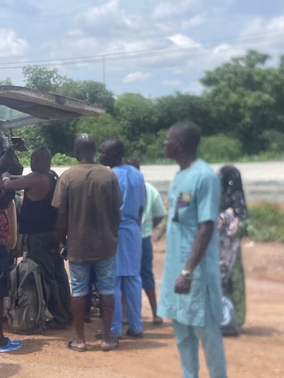 The image shows a group of people standing outdoors on a dirt road. In the foreground, a man in a light blue traditional garment is looking to the left. He is wearing a watch on his left wrist. To his right, a woman in a patterned headscarf and dress is visible. Behind the man in blue, a man in a brown t-shirt and denim shorts is facing away from the camera. To his left, another man in a blue traditional garment is also visible. In the far left of the image, a person wearing a black tank top and camouflage pants is partially visible, with a large backpack next to them. Overhead, a wooden structure is partially visible. The background consists of trees and a cloudy sky. The scene appears to be in a rural or semi-rural area of Osu, Nigeria.