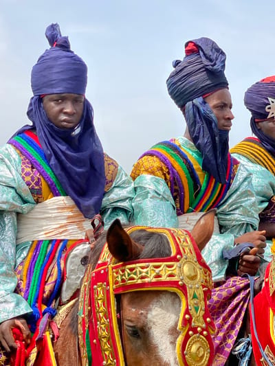 The image depicts three men on horseback in Kafanchan A, Nigeria. Each man wears a dark blue turban, a light blue, patterned robe, and a colorful rope around their chest. The horses are elaborately decorated with red and gold headgear. The background is a pale sky.