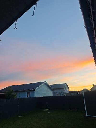 The image depicts a backyard scene at dusk, with a vibrant sunset painting the sky in hues of pink, orange, and blue. The foreground features a dark, textured fence and a neatly mowed green lawn. Beyond the fence, two houses with grey roofs are visible, one on the left with light blue siding and a single window, and another on the right with brick and siding construction. The sky above is a gradient, transitioning from a clear blue at the top to the warm colors of the sunset closer to the horizon. The image appears to be taken from within a property, with the dark eaves of a structure framing the top edges of the photograph. The location is identified as Kostomarovo, Russia.