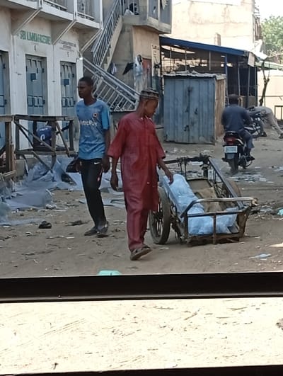 The scene depicts an unpaved street or market area in Kofar Ruwa, Nigeria, under daylight conditions. The foreground features a dark horizontal bar and a light, textured surface. The ground is primarily dirt or sand, littered with numerous white plastic bags and other debris.
In the mid-ground, two young males are prominent. On the right, a male wearing a dark red long-sleeved tunic and a kufi (cap) walks left-to-right in sandals, looking downwards. Immediately to his left is a two-wheeled metal pushcart containing multiple clear plastic bags filled with what appear to be water sachets. To the left of the first male, a second young male walks left, wearing a light blue short-sleeved shirt with "MetroPay" printed on it, dark trousers, and dark slip-on footwear.
In the background, a third male is visible on the right, seen from behind, riding a dark motorcycle. Further right and partially obscured, a fourth individual in light-colored clothing is bent over. The background structures include a multi-story light-colored building on the left with several light blue closed doors and an external metal staircase. Text partially visible above the doors includes "SBI ALU..." and "AL-IMAM..." To the right, another building features a blue corrugated roof extension and a dark metal shed. Various wooden market stall frames and white plastic sheeting are also present throughout the scene.