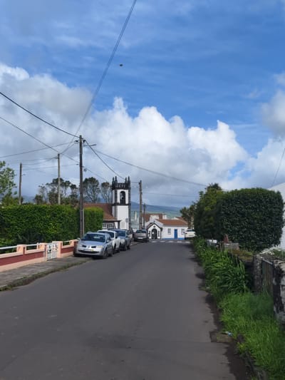 A daytime view looking down a narrow asphalt road in Ponta Delgada, Portugal. The road is lined with parked cars on the left, and a mix of overgrown greenery and stone walls on both sides. A small church with a white facade and black trim on its bell tower is visible further down the road. Blue and white buildings are seen on the right. Power lines are strung across the sky, which is partially covered by large, fluffy white clouds. In the distance, a mountain is visible.