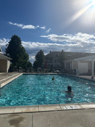 In Allen Township, United States of America, a rectangular swimming pool with crystal clear blue water is pictured under a bright, sunny sky with scattered clouds. Two individuals are present in the water; one is further away, appearing to be a child with a green ball, and another is closer to the foreground, a person with dark hair and wearing swim trunks. The pool is surrounded by a concrete deck, with lounge chairs and umbrellas visible in the background. Adjacent to the pool is a building with light-colored siding and a dark roof, and further in the background are more residential buildings and lush green trees. A depth marker indicating "5' 0\"" and a "NO DIVING" sign are visible on the pool's edge. The overall scene depicts a recreational area on a pleasant day.