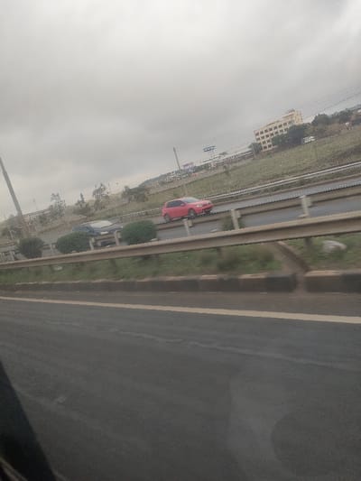 The image displays an overcast daytime scene of a multi-lane highway in the Gatongora ward, Kenya. In the immediate foreground, a dark asphalt road surface with a yellow edge line is visible on the right. A three-bar metal guardrail runs horizontally across the mid-section, separating the foreground from active traffic lanes.
Behind the guardrail, two vehicles are in motion, traveling from right to left. A dark-colored hatchback or sedan is partially visible in the left lane, while a red hatchback, identified as a Volkswagen Golf, is fully visible in the right lane. These vehicles are on a paved road surface, with a grassy median containing several spherical green bushes separating the lanes.
In the background, an elevated grassy embankment with sparse trees and utility poles is present. A multi-story, light-colored building with visible windows stands prominently on the upper right of this embankment. A white truck or van is discernible on a higher road section or parking area near the building. Further in the distance, indistinct blue signage or a billboard is partially visible above the embankment. The sky is uniformly overcast, indicating dense cloud cover. No pedestrians are visible.