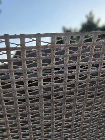 This media file features a close-up, angled view of a damaged, perforated metal grate. The grate is predominantly gray and shows signs of wear and tear, with small, multicolored flecks embedded in its surface, possibly from paint or debris. The perforations are rectangular with diagonal lines connecting the corners, creating a lattice-like pattern. Fine strands, likely cobwebs, are visible across the surface of the grate.

In the background, to the left of the grate, a blurry, out-of-focus scene is visible. This includes what appears to be greenery, possibly trees or bushes, and a light blue sky. The overall lighting suggests it is daytime, but the weather conditions are indistinct due to the blur.

The image captures a static scene, with no discernible activity or event taking place. The focus is entirely on the texture and pattern of the damaged grate. The context of Nova Kakhovka, Ukraine, suggests a setting that may have been affected by conflict or damage. There is no visible text in the image.