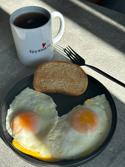 An overhead shot captures a simple breakfast setup on a textured, light-colored surface, likely a table, in Tomilino, Russia. The scene is bathed in natural sunlight, casting sharp, diagonal shadows across the surface, indicating a clear, sunny morning or late afternoon.

The main subjects are a black, round plate holding two perfectly cooked sunny-side-up fried eggs with bright orange yolks and a slice of plain, lightly toasted bread. Adjacent to the plate, on the left, is a white ceramic mug, filled with a dark beverage, likely coffee or tea. The mug features Cyrillic text printed in black, which reads "Кружка" (Kruzhka), meaning "Mug," followed by a small red heart icon. A black fork is placed diagonally in the upper part of the image, resting on the table surface, partially within the shadows. The composition suggests a peaceful, individual meal.