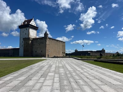 The image shows a wide view of Narva Castle in Narva, Estonia, on a sunny day with scattered clouds. In the foreground, a large paved area with gray stone tiles extends towards the castle. To the left of the paved area is a neatly mowed green lawn. The castle itself is a prominent feature, with a tall, white tower on the left, and a long, stone building with smaller turrets extending to the right. The castle walls are constructed of light brown stone. A dark flag flies from a pole on top of one of the turrets in the distance. The sky is a vibrant blue with fluffy white clouds. In the background, more of the castle's extensive fortifications can be seen. A lone figure is visible standing in front of the distant wall, appearing to look towards the castle. The overall scene depicts a historic landmark under a bright, clear sky, with a clean and well-maintained public space in front of it.