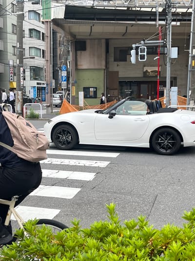 In Taito, Japan, on a city street with a pedestrian crossing, a white convertible car is parked on the right side of the road. A person wearing dark clothing and a light brown backpack is on the left, seated on a bicycle, with their back to the viewer. The car has black wheels and the top is down. In the background, buildings are visible, including one with signage that reads "BASE CAMP COFFEE." Traffic lights and street signs are also present. The weather appears to be overcast.
