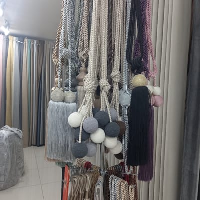 The image depicts a retail display of curtain tiebacks and tassels in a store. The tiebacks are made of braided cords in various colors including beige, grey, silver, black, and purple. Many of them are adorned with decorative elements such as knotted cords, tassels made of thin strands, and spherical ornaments covered in beads or woven with yarn. In the background, several draped curtains in muted tones of pink, yellow, grey, beige, and green are visible. The floor is tiled with large square tiles. The overall setting suggests a fabric or home decor store. No people are present in the image. The image was taken in Briceni, Moldova.