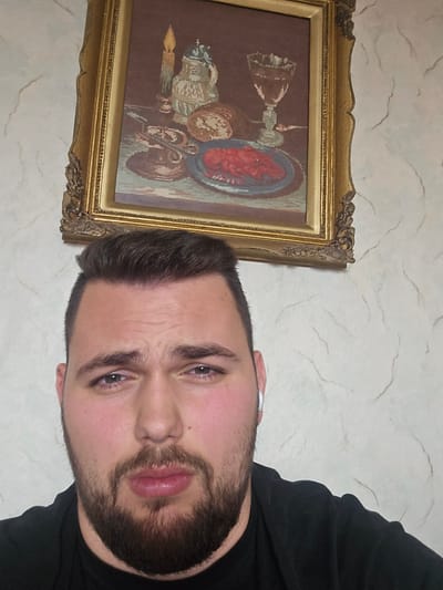 An indoor photograph features an adult male occupying the lower foreground, directly facing the camera. He possesses dark brown hair, a full dark brown beard, and is dressed in a black shirt. A white earbud is visible in his right ear. His expression is characterized by slightly furrowed eyebrows and a neutral mouth. Positioned on the wall behind him, slightly to his left, is a rectangular framed artwork with an ornate gold frame. The artwork, appearing to be a needlepoint or cross-stitch, depicts a still life featuring a lit candle, a decorative pitcher, a loaf of bread, a plate with a red crustacean (lobster), and a stemmed glass. The wall itself is light-colored with subtle, irregular, wavy patterns. The location depicted is Han Krum, Bulgaria.