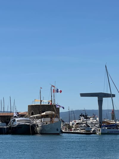 This is a medium shot of a marina in Saint-Tropez, France on a clear, sunny day. The foreground is dominated by the blue water of the harbor, with several boats docked. In the middle ground, a line of moored yachts of various sizes are visible. To the left, a large, dark-colored motor yacht with a sleek design is prominent. Adjacent to it is a larger, light-colored boat with a classic design, featuring wooden accents and a multi-story cabin. Behind these boats, a circular, stone tower with a red-tiled roof stands. Several flagpoles adorn the tower and boats, flying various national flags, including the French flag. Further to the right, a large white gantry crane stands, with "SAINT-TROPEZ" written on its side. Beyond the marina, a range of green hills forms the background, under a bright blue sky with a few faint clouds and a distant seagull. The overall scene depicts a bustling harbor with a mix of modern and classic vessels.