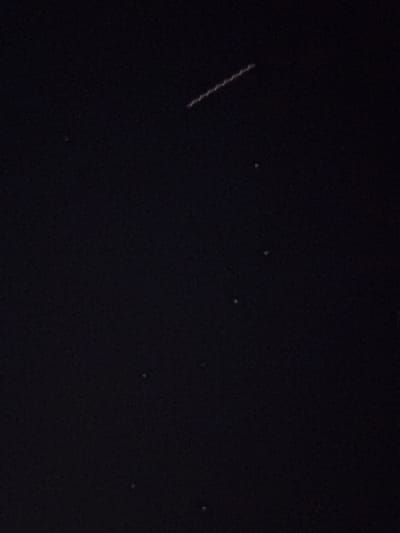The image is a nighttime photograph of the sky above Shkorpilovtsi, Bulgaria. The sky is dark, with a few scattered stars visible. A trail of lights, likely from a satellite or aircraft, is visible diagonally across the upper portion of the image. The trail is composed of small, distinct lights. No other objects, people, or structures are discernible in the frame. The environment appears to be an outdoor, unlit area, consistent with a clear night sky.
