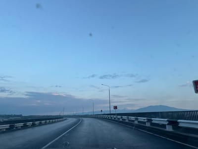 The image depicts a highway bridge at dusk, with a clear sky overhead and a hint of clouds on the horizon. The road is wet, suggesting recent rain, and illuminated by streetlights. In the distance, mountains are visible against the twilight sky. The bridge is equipped with a metal guardrail that has black and white reflective markings. The scene appears to be located in or near the city of Arzni, Armenia, given the characteristic landscape and potentially the architecture in the background. There are no people or vehicles visible in the image.