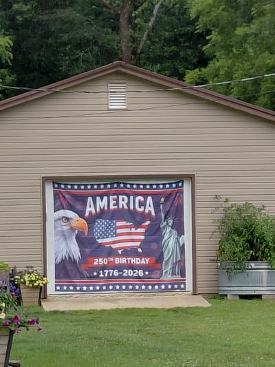 The image shows a banner displayed on a garage door. The banner features a bald eagle on the left, a map of the United States with the American flag superimposed on it in the center, and the Statue of Liberty on the right. Text on the banner reads "AMERICA", "250TH BIRTHDAY", and "1776-2026". The garage is clad in beige horizontal siding and is topped with a brown roof. A ventilation grate is visible near the peak of the garage. The scene is outdoors, with green trees in the background and a grassy lawn in the foreground. To the right of the garage, a large galvanized metal container filled with green plants is visible. The location is not specified in the image or its metadata.