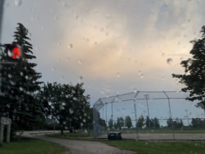 The image captures an outdoor scene through a window covered in raindrops. In the foreground to the left, a traffic light is visible with a red light illuminated. Behind the traffic light, a tall evergreen tree stands prominently. Further to the right, a baseball field with a chain-link fence and lights is present. Trees and grassy areas surround the field. The sky is overcast with some lighter patches suggesting a cloudy day, possibly during dusk or dawn. The presence of raindrops indicates it is raining. The overall mood is subdued due to the weather conditions. There is no visible text in the image.
