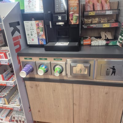 The image displays a close-up view of a coffee machine and surrounding retail display in Bralin, Poland. In the foreground, a modern coffee machine with a black finish is prominent. To the left of the coffee machine, a large white and red "K" logo is visible on a display stand, partially obscuring some of the merchandise. Below the coffee machine, there are several dispensers for cups and a trash receptacle. One dispenser, labeled "duża" (large), has a purple cup inserted. Next to it, labeled "mała" (small), a green and blue patterned cup is present. A third dispenser, labeled "espresso," contains a stack of green cups. To the right of these dispensers, a stainless steel slot holds a folded beige paper, possibly a lid or sleeve, with the text "dobregodnia!" (have a nice day!) and "żabka" (frog, likely the brand name). The final slot on the right is a trash receptacle with a silhouette of a person throwing an object into a bin. The background shows shelves stocked with various food items, including bags of chips labeled "NATCh," individually wrapped muffins, and boxes of Kinder chocolate products. The overall environment appears to be a self-service area within a convenience store or gas station.