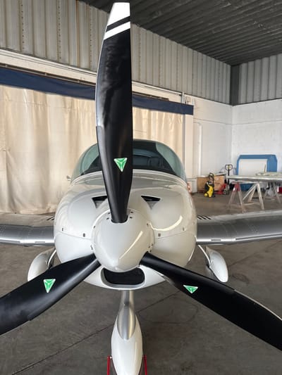 A white, single-engine, three-blade propeller aircraft is centrally positioned in a hangar at Toussus-le-Noble, France, viewed from a slightly elevated frontal perspective. The aircraft features a glossy white fuselage and engine cowling, with silver-colored wings exhibiting a riveted surface. Its three propeller blades are black, each marked with two horizontal white stripes near the tip and a green triangular emblem outlined in white near the root; minor smudges are visible on the blades. A clear cockpit canopy reflects internal lighting. The aircraft's nose landing gear is visible, resting on a red ground support stand. The hangar environment includes light gray corrugated metal walls and a dark gray concrete floor. In the left and central background, a light-colored fabric curtain with a dark blue header delineates a partitioned area. The right background reveals a workshop zone containing a blue-covered object, a wooden workbench on sawhorses with a light-colored component, a yellow Kärcher pressure washer, and assorted boxes and equipment. No individuals are present in the scene.