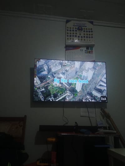 A wall-mounted TV displays a bird's-eye view of a city with text overlay reading "By His own hand" and "SoA" logo. Above the TV hangs a calendar, showing December 12, and a larger calendar with circular images. Below, a cluttered desk holds electronics, cables, and a guitar rests against the wall. To the left, a chair back is visible in the dim lighting.