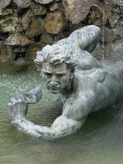 The image is a close-up, eye-level, vertical shot of a weathered, green-patinated bronze statue of a male figure. The statue is partially submerged in murky, green water, with water cascading down from a rocky, naturalistic background. The figure is depicted in a dynamic pose, seemingly emerging from or interacting with the water. The musculature of the torso and arm is prominent, rendered with detail despite the patination. The figure's face, with its furrowed brow and intense gaze, is a focal point. In its left hand, the figure holds a mask-like object with a circular opening. The water creates a spray and ripples around the statue, adding a sense of movement.

This statue is part of the Grelin Fountain, located in the Place de la Comédie in Bordeaux, France.