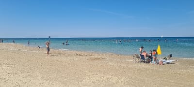 The image shows a sandy beach with people sunbathing and swimming in the water.  The beach is located in the city of  Mihmoret, Israel, at coordinates (latitude: 32.408, longitude: 34.868). The weather is sunny with clear blue skies.  In the distance, there is a group of people standing on paddle boards in the water. Several people are scattered on the beach, including a group of four people sitting under a yellow beach umbrella. One man is standing on the beach, facing away from the camera, holding a surfboard.