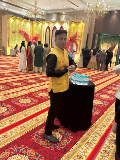 A man stands on an ornate red and gold patterned carpet, wearing a black shirt, black pants, and a yellow vest. A black table draped with bottles of water is next to him. The background shows a large hall with gold-decorated walls and chandeliers. Several people in traditional attire are milling around, some lined up as if waiting. The scene likely depicts an event or gathering, potentially a wedding or celebration in an opulent setting.
