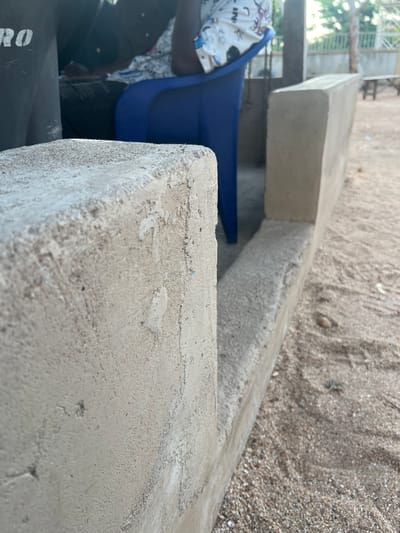 An outdoor, low-angle close-up shot shows a section of a sandy area with two concrete structures in the foreground. The structure on the left is a roughly finished concrete post, angled towards the viewer. To its right is a longer, more smoothly finished concrete wall or ledge. The sandy ground is visible between and beyond these structures, with small pebbles and debris scattered throughout. In the background, partially visible, are two individuals seated on chairs. One person is wearing a blue shirt and the other is wearing a white patterned top. A dark, possibly black object with white lettering is on the far left. Beyond them, a metal fence and trees are visible, suggesting a semi-urban or suburban environment. The lighting indicates it is daytime, with shadows cast on the ground. The scene appears to be in Mayo Dasa, Nigeria, an area characterized by its sandy terrain and common use of concrete structures for everyday purposes.