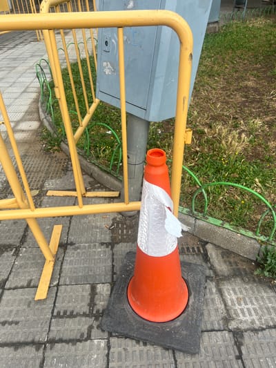 The image captures an outdoor scene, predominantly featuring an orange traffic cone and a yellow metal barrier on a paved surface next to a grassy area. The photograph is oriented with a significant tilt, presenting the ground plane running diagonally from the top right to the bottom left.
On the right side of the frame, an orange traffic cone with a sturdy black base stands on textured concrete paving slabs. A white reflective band, featuring a honeycomb pattern, is wrapped around the cone's upper section but is notably torn and peeling in several places. The cone's top opening reveals its orange interior.
To the left of the cone, a bright yellow metal barrier extends across a significant portion of the image. This barrier, composed of multiple horizontal rungs and vertical supports, shows signs of wear, including scuffs and dirt. Behind the barrier, a gray utility box or cabinet is partially visible.
Further to the left and in the background, a patch of green grass is seen, interspersed with dry weeds and small yellow flowers. A bright green hose or cable snakes irregularly across this grassy area. The ground around the cone consists of gray concrete pavers, some of which appear damp or stained. Scattered cigarette butts and fragments of yellow material, possibly broken pieces of the barrier, lie on the pavement.
Visible text on the black base of the traffic cone includes "TRAFFIC CONE CLASS S2 C2 V2", "EN 13422", and "A1 CONO7500". The scene is illuminated by natural daylight, suggesting it was taken during the day. No individuals are present, and the setup of the cone and barrier indicates a temporary obstruction or designated area.