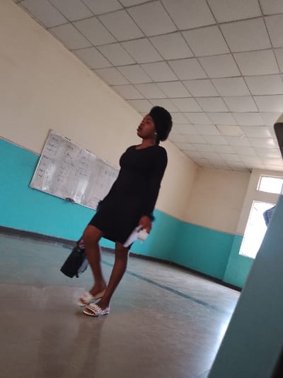 A woman is walking in what appears to be a school hallway. She is wearing a black dress, and sandals. She is carrying a black purse. A whiteboard with equations is visible on the wall behind her. The walls are cream-colored with a blue stripe. The ceiling is a grid of square panels. The location is Umueshi, Nigeria.