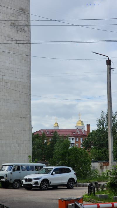 The image shows a street scene in Smolensk, Russia. A brick building with golden domes and crosses is visible in the background, suggesting the presence of a church or religious institution. In the foreground, a tall concrete or stone wall runs horizontally across the lower portion of the image. Power lines and a utility pole are situated on the left side, extending vertically toward the sky. A white BMW is parked on the right side of the image, partially blocking the view of a light grey van parked behind it. Both vehicles are facing the same direction. The weather appears cloudy. A striped barrier and a green tree are visible on the right side behind the car.