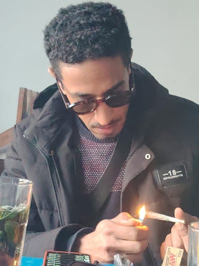 A man with dark curly hair and sunglasses sits indoors. He wears a dark jacket over a patterned sweater. He is lighting what appears to be a hand-rolled cigarette with a lighter. A pack of cigarette rolling papers is on the table, next to a glass of a dark liquid with green sprigs. He looks down at the cigarette, concentrating on lighting it.