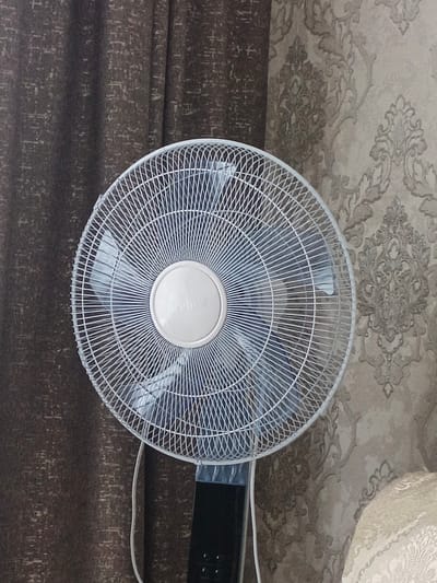 A white oscillating fan is positioned in the foreground, occupying the majority of the frame. The fan has a silver circular center with faint text on it. Behind the fan, on the left, is a dark brown curtain with a textured pattern. To the right of the fan, the background consists of a patterned wallpaper in beige and taupe tones, featuring ornate floral motifs. The lighting appears to be indoor and diffused, casting soft shadows. The image does not provide any information that would allow for its location to be identified as Sukhodol, Russia, or any other specific city.