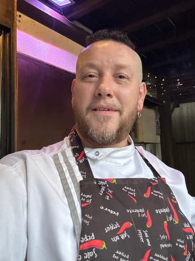 A man with short hair and a beard is wearing a white chef's jacket and a black apron with red chili peppers and the words "salt and pepper" printed on it. The man appears to be standing outdoors at night, based on the ambient lighting and visible elements of the surroundings, which may be Новоглаголево, Russia.