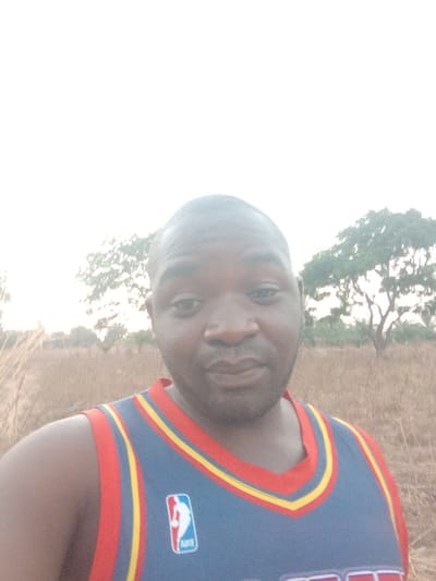 A male individual with a dark complexion, shaved head, and short facial hair is depicted in a chest-up, forward-facing shot. He is wearing a blue sleeveless basketball jersey with red and yellow trim, featuring the white NBA logo and "BHV" text on the left chest. The subject maintains direct eye contact with the camera, displaying a neutral to slight smiling expression. The background, set in Gudi, Nigeria, consists of an open, outdoor area with dry, yellowish-brown ground covered by sparse vegetation. Two trees, one prominent with green foliage to the right and another partially visible to the left, are present against an overexposed, light-colored sky.