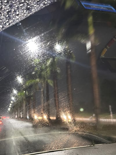The image, taken from inside a vehicle, depicts a street scene at night in Gollapalle Khurd, India, during rainfall. The windshield is covered in water droplets, creating a blurry and distorted view of the surroundings. Rows of palm trees are visible along the street, illuminated by streetlights. The wet asphalt reflects the lights, and car headlights can be seen in the distance. The overall atmosphere is dark and moody, characteristic of a rainy night.