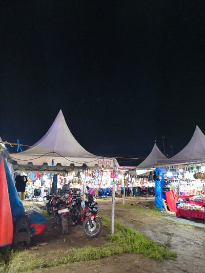 The image depicts an outdoor night market operating in An Khe Ward, Vietnam. The scene is illuminated by artificial light emanating from within and around several white, pyramid-roofed temporary tents erected on an uneven dirt ground with sparse patches of grass. The sky overhead is dark and featureless.
Under the central and leftmost tents, a line of approximately four motorcycles, primarily red and dark-colored, are parked. A red Yamaha scooter is prominent in the mid-ground. Each tent houses market stalls displaying various goods; items resembling clothing, accessories, and general merchandise are visible on racks and tables. Blue-cased string lights hang beneath the tent canopies. A sign with the word "DADI" is visible above a central stall. On the far right, another stall displays merchandise on tables draped with red fabric, adjacent to a blue tarp. No distinct individuals or specific actions are discernible within the frame.