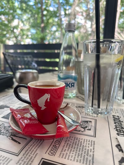 The image shows a table set for a drink, possibly at an outdoor cafe. In the foreground, a red mug with a white silhouette of a bird sits on a saucer. Beside the mug, there are two small red packets, one labeled "Julius Meinl CARAMEL BISCUIT." A spoon rests on the saucer. Behind the mug, a bottle of water is visible, with "КЯ" printed on the label. Further back, a tall glass contains a clear liquid and a black straw, with a slice of lemon near the top. The table is covered with a newspaper-like material, featuring headlines and text in a gothic font, including "GOSSIP COLU" and "spape." The background is blurred, showing greenery and what appears to be dappled sunlight, suggesting an outdoor setting on a bright day. The overall impression is one of a relaxed moment, perhaps enjoying coffee or a beverage outdoors. The text "Petrich, Bulgaria" provides a specific location context.