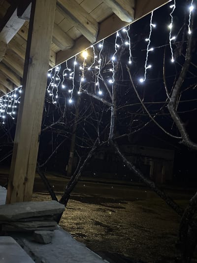 The image depicts an exterior night scene in the city of Касапите, Bulgaria, characterized by cold, wet conditions. In the foreground on the left, a portion of a wooden architectural structure, likely an overhang or porch, features light-colored timber beams and a prominent vertical support post. White LED icicle string lights are actively illuminated, suspended from the edge of this structure. Below the wooden post, a small stack of flat, irregular grey stones rests on a dark, indeterminate surface. The mid-ground is dominated by several bare, dark deciduous tree branches, appearing wet and reflecting light, suggesting moisture or ice. Small, bright specks in the air indicate falling precipitation, potentially light snow or freezing rain. The background consists of a dark, wet, reflective paved surface, consistent with a road or pathway. A single amber-colored streetlamp is visible in the upper mid-ground, casting a localized glow. Distant, indistinct dark structures, likely buildings, and a utility pole are visible in the deep background with limited illumination. No individuals or animals are present in the scene.