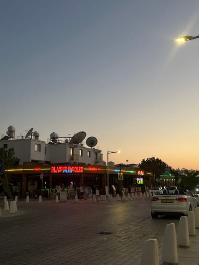 This media file captures a street scene in Peyia, Cyprus, at dusk. The sky is a gradient of soft orange and pale blue, suggesting the sunset. The foreground is dominated by a paved walkway lined with white bollards.
On the right side of the image, a white Audi car is parked, displaying a license plate that reads "KZN 110". Beyond the car, a row of buildings with multiple satellite dishes on their roofs is visible. One building prominently features a bright neon sign that reads "BLAZING SADDLES PUB" in red lettering, with smaller signs for "COCKTAIL BAR" and "CAFE" above and below. The facade of the pub appears to be illuminated by yellow and green lights.
In the background, trees and more buildings can be seen. There are glimpses of people walking and congregating outside the establishments. The overall atmosphere is relaxed and hints at an evening social setting, perhaps as bars and cafes are opening for the night. A street lamp with a bright yellow light is visible in the upper left corner of the image. The scene is illuminated by a combination of natural twilight and artificial lights from the businesses.