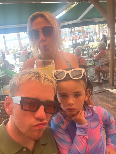 The media file shows a group of people at an outdoor cafe, likely in Benidorm, Spain, given the text "Joke board Benidorm" visible in the background. The image is oriented vertically, with the subjects filling the foreground. In the center of the frame, a young girl with dark hair wears white-rimmed sunglasses and a tie-dye top. To her left, a woman with blonde hair and large brown sunglasses also wears sunglasses. Her mouth is slightly open, and she appears to be speaking or reacting. To the right of the girl, a man with short blonde hair, a light beard, and dark-rimmed sunglasses is partially visible, wearing a green shirt. The group appears to be at a table, with a drink containing a lemon slice in the foreground.

In the background, other patrons are visible at tables and chairs. The setting suggests a sunny day, with bright light casting shadows. The overall atmosphere seems relaxed and casual, indicative of a vacation or leisure activity. The text "Joke board Benidorm" is clearly legible on a sign in the background, confirming the location. Other people and beach umbrellas are visible further in the background, suggesting a coastal or tourist area. The close-up nature of the foreground subjects, particularly the child and the woman, hints at a selfie or a candid shot taken by one of the individuals.