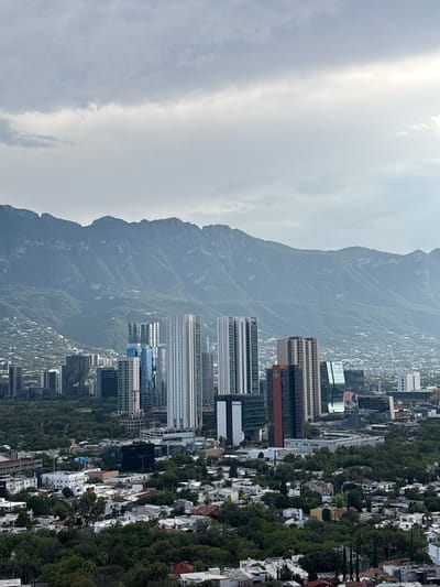 The image captures a panoramic view of San Pedro Garza García, Mexico, under a cloudy sky. In the foreground, a dense canopy of green trees partially obscures a residential area with houses featuring red-tiled roofs and white walls. Moving further back, a collection of modern skyscrapers, characterized by glass facades and varying heights, dominate the urban landscape. The tallest buildings are a cluster of white and glass towers. Beyond the city, a prominent mountain range forms the backdrop, its slopes covered in lush greenery and dotted with scattered structures. The overall scene depicts a harmonious blend of natural beauty and urban development.