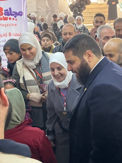 An indoor scene in Kafr Batna, Syria, depicts a crowded event or gathering. In the immediate foreground, a man with a dark beard, wearing a dark blue suit jacket over a light collared shirt, is seen in profile on the right, looking down and to his left with a smile. Adjacent to him, a woman wearing a white hijab and a grey coat with a red lanyard and a dark identification badge around her neck is smiling and looking towards the bearded man. To her left, another woman with a beige hijab, a grey vest, a beige and striped scarf, and light-colored fingerless gloves is smiling and looking downwards. A small brown cross-body bag is visible across her chest. A young boy in a dark beanie is positioned to her left, looking down. In the mid-ground, a diverse crowd of individuals, including several men and women in hijabs, is present. A young boy with dark markings on his cheek, wearing a yellow jacket, is visible. Another woman in a white hijab and a black and white plaid shirt stands further back. In the background, a large white banner with red Arabic text, including the word "Magazine" and "8 كانون" (8 January), is visible on the left. To the right, a set of light-colored stairs adorned with black balloons ascends. The overall scene suggests a public event with positive interactions among attendees.