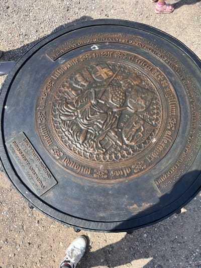 This is a medium, high-angle, outdoor, close-up, full shot of a large, circular, bronze plaque embedded in the ground, likely made of gravel. The plaque features an embossed central design of a knight on horseback, possibly Saint George slaying a dragon, surrounded by a decorative border with what appears to be Lithuanian text and Braille. Some of the text is partially obscured by shadow. On the left side of the plaque, a separate rectangular plaque with more text and Braille is affixed. The surrounding ground is covered in small, light-colored gravel and pebbles. In the upper right corner, the feet of a child wearing pink sandals are visible. In the lower portion of the image, a white sneaker with red and blue accents is positioned near the plaque. The overall lighting suggests a sunny day, with distinct shadows cast by the plaque and the surrounding objects. This plaque is located at the site of the Trakai Castle in Trakai, Lithuania, marking the "Footprint of Vytautas the Great."