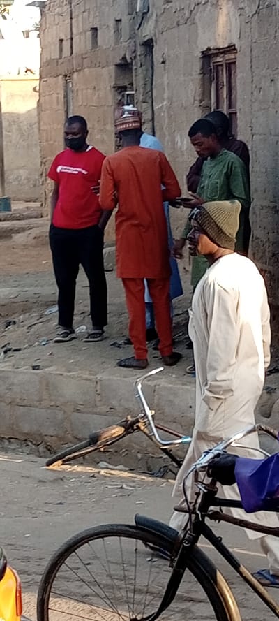 The image depicts a street scene in Kabuga, Nigeria, featuring six adult males and two bicycles near a dilapidated building.

In the foreground, two bicycles are present. On the lower left, part of a yellow vehicle with a reflector is visible. Adjacent to it, the front wheel and handlebars of an upright bicycle are seen, with a purple fabric draped over the handlebars. To the right, a second bicycle lies horizontally on the dusty ground, partially obscured by a low concrete curb.

On a raised concrete platform or steps behind the bicycles, five men are gathered. From left to right:
1.  A man wearing a red t-shirt, black trousers, and a black face mask stands with his hands in his pockets, facing right.
2.  Next to him, a man in an orange long-sleeved traditional tunic and matching trousers, wearing a patterned cap, stands with his back largely to the viewer, facing right. He wears dark slippers.
3.  Partially obscured behind the man in orange is a third man in a light blue traditional garment.
4.  Further right, a man in a green long-sleeved traditional shirt is looking down at a mobile phone he holds in his hands.
5.  Partially visible behind the man in green is another individual in a dark garment.

In the right foreground, slightly separated from the group on the platform, a sixth man is seen in profile, facing left. He wears a light-colored (possibly off-white or cream) long traditional tunic and a brown and olive green knitted beanie. He has a short beard.

The background consists of a weathered, light-colored building constructed of concrete blocks or mud bricks, featuring several recessed windows, some appearing boarded up or partially filled. The ground is unpaved, dusty, and uneven, with visible debris. The scene is illuminated by natural daylight.